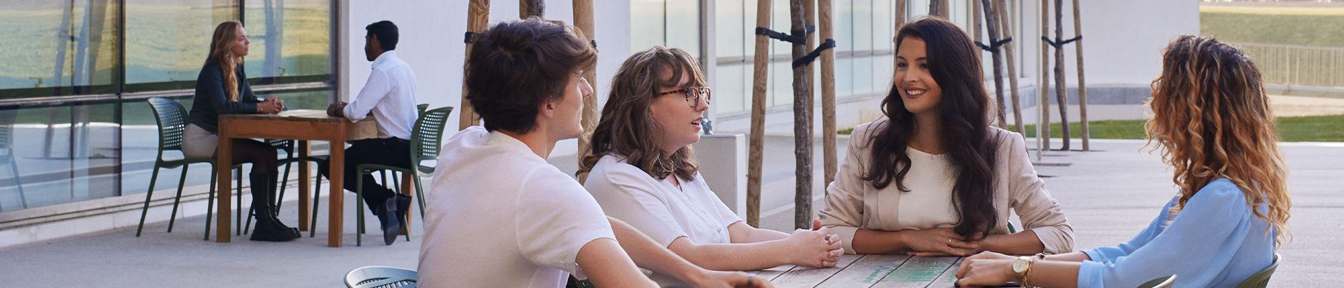 A group of five people engaged in conversation at an outdoor table, with two others sitting at a separate table in the background.