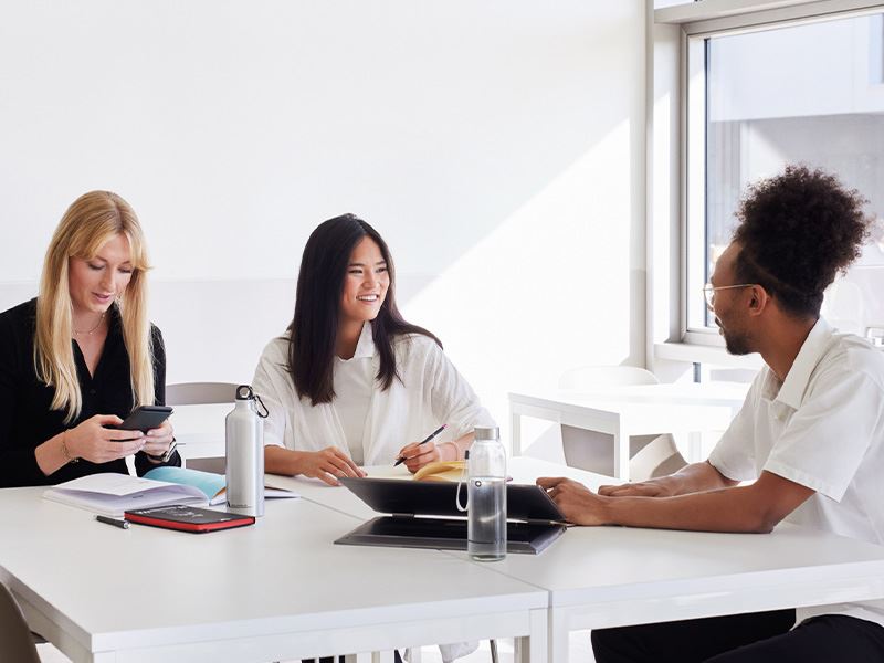 Three business professionals engaged in discussion at a table, each using a laptop for collaboration and communication.