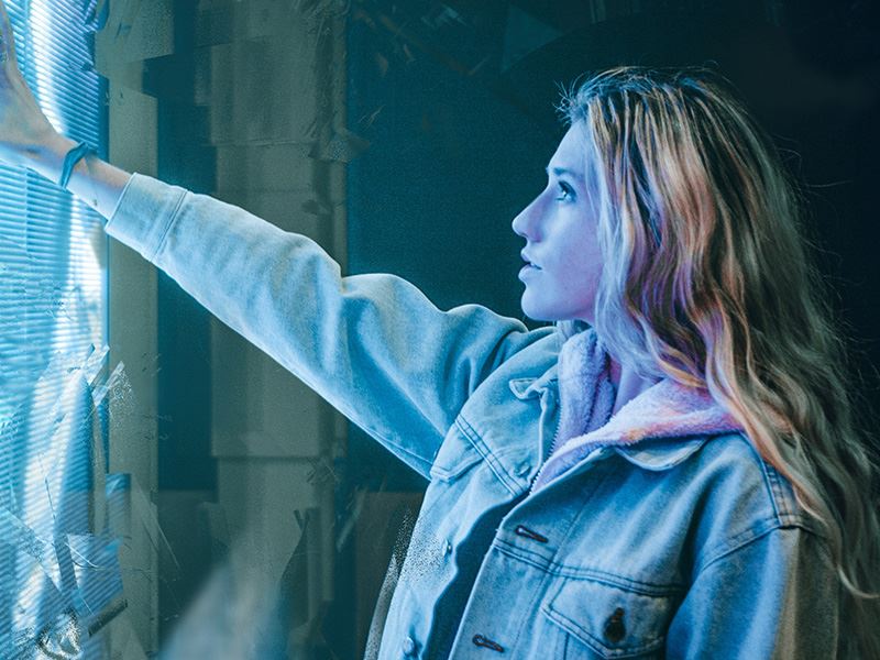 A young woman with long, wavy hair is reaching out towards a screen or surface, illuminated with a blue light. 